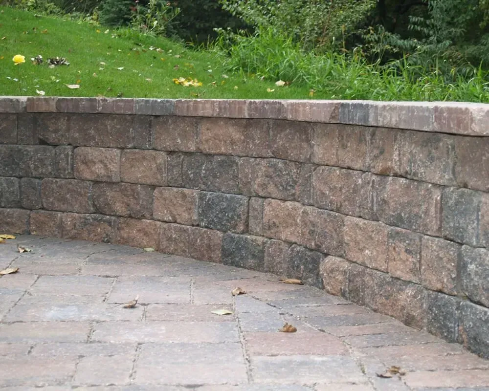 Curved brick retaining wall with a paved patio in front and green grass and foliage behind.