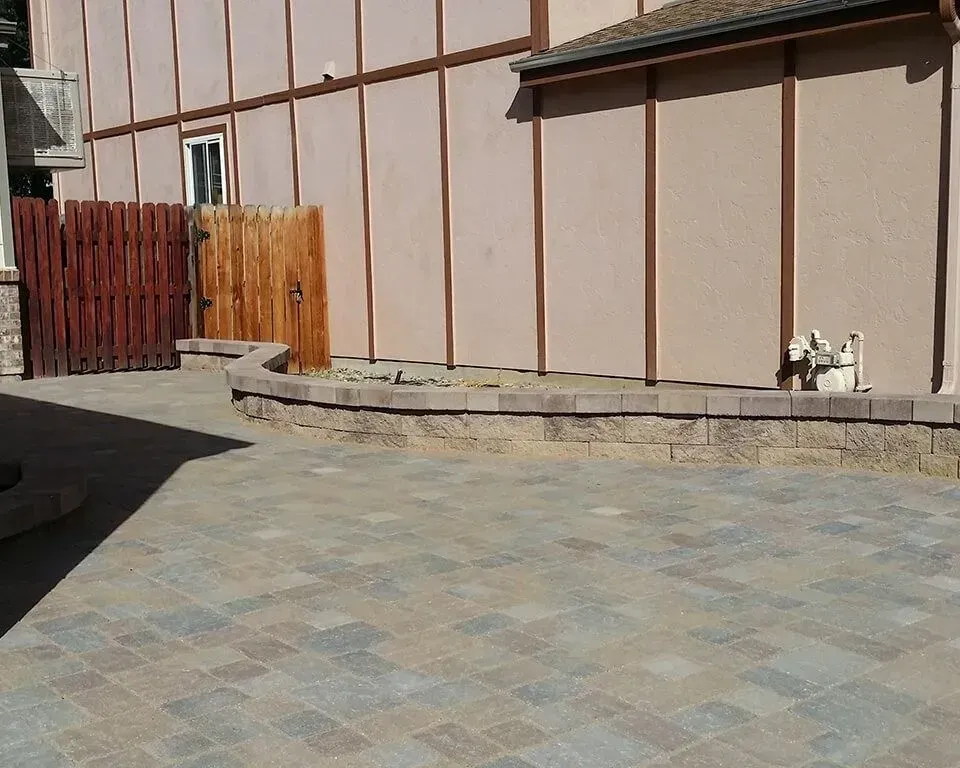 Paver patio with low retaining wall and wood fence against a beige building.