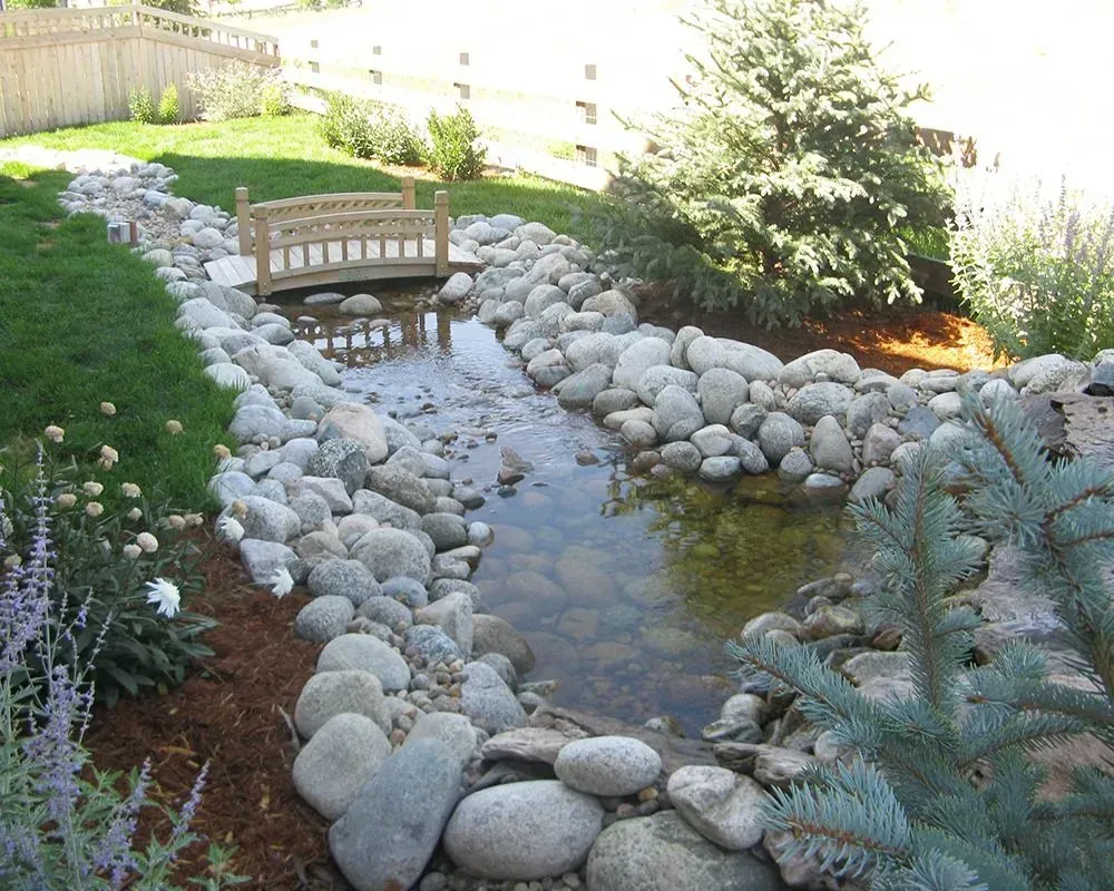 A decorative backyard stream with a small wooden bridge and rocks along the banks.