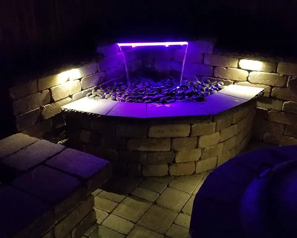 Nighttime outdoor water fountain with purple light and surrounding stonework.