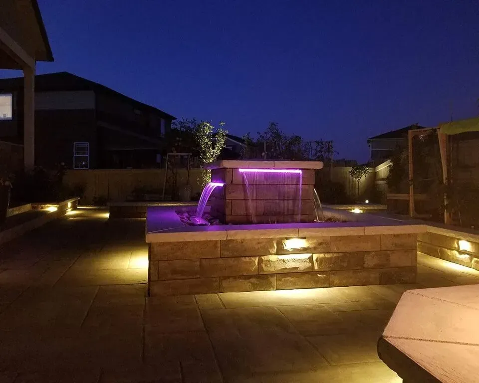 Nighttime outdoor scene with lit waterfall and landscaping. Purple lights illuminate the water, brown stone walls.