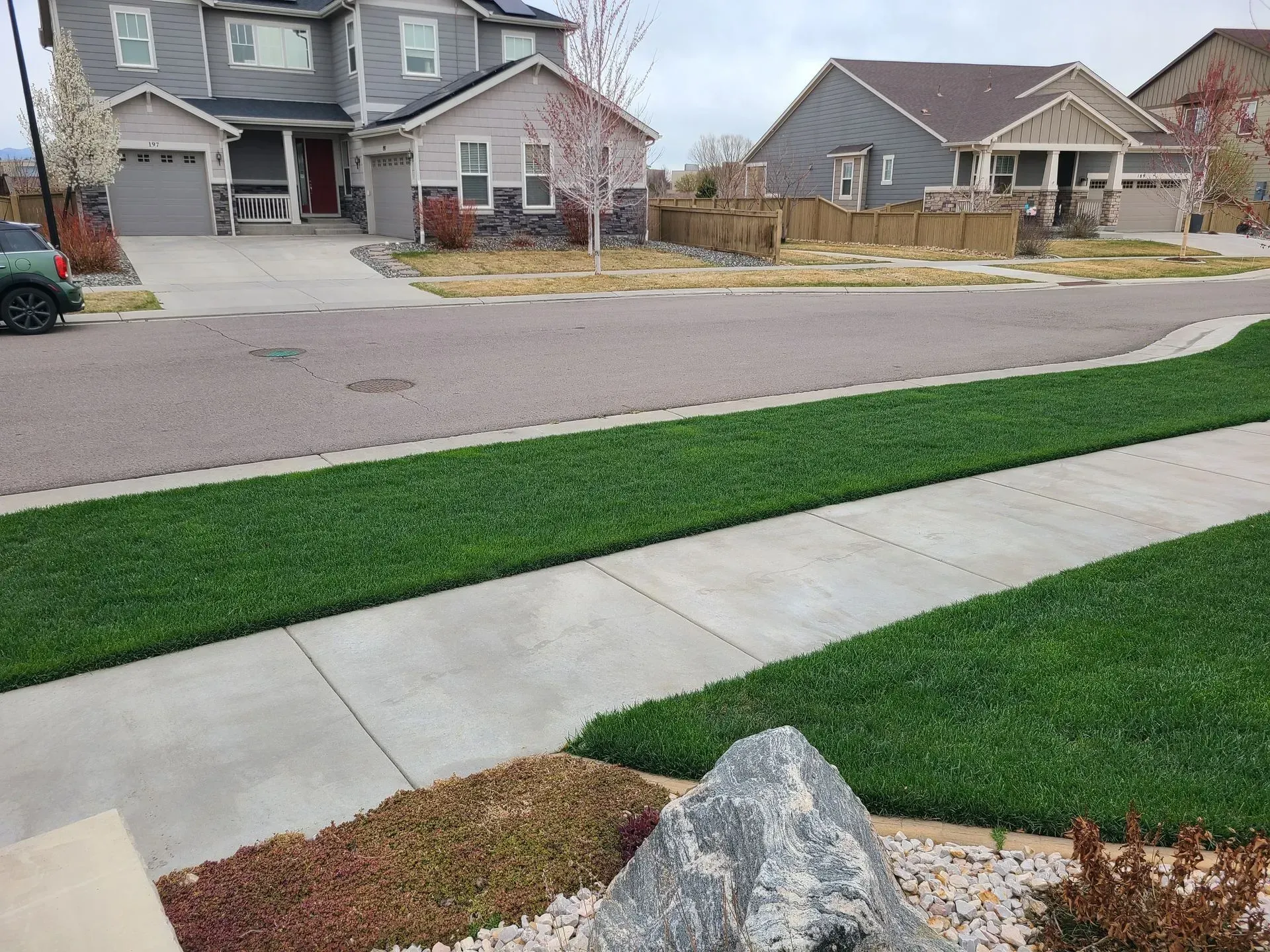 Residential street scene: houses with driveways, green lawns, sidewalk, and roadway. Cloudy sky.