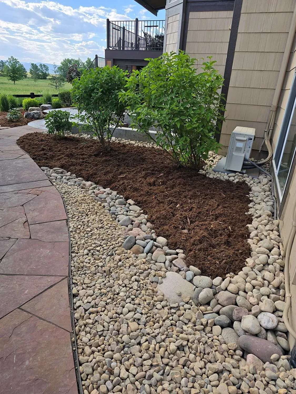 Landscaped garden bed with mulch, river rock, and shrubs, next to a building and paved walkway.