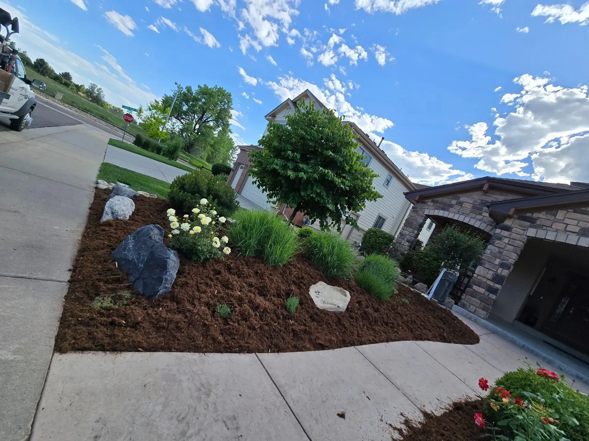 Landscaped flower bed with mulch, large rocks, and various green plants in front of a house.
