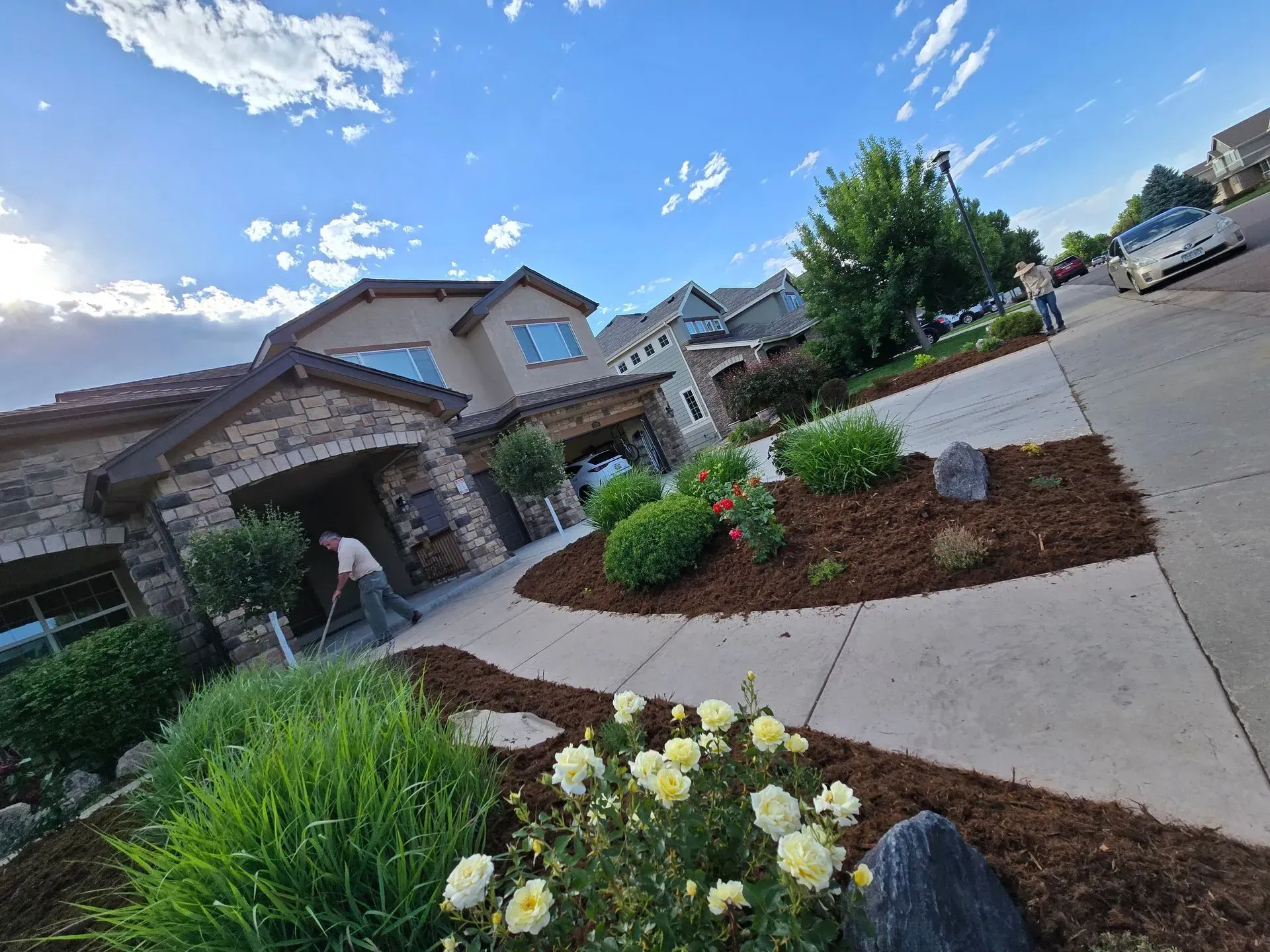 House with stone facade, driveway, and landscaping. A person rakes mulch. Sunny day with blue sky.