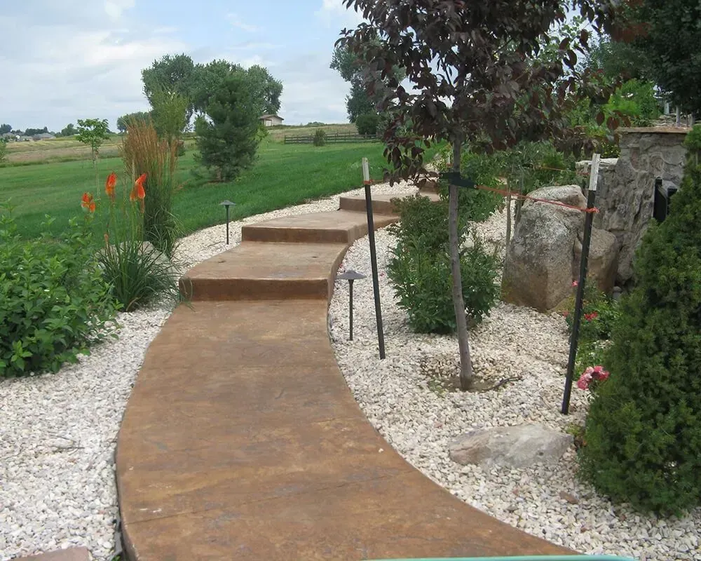 Brown curved concrete pathway with steps, lined with white stones and landscaping.