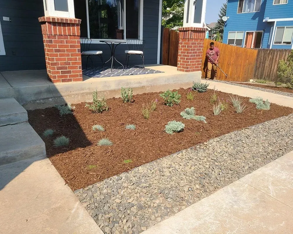 Front yard garden bed with plants, wood mulch, and gravel border. A person stands nearby.