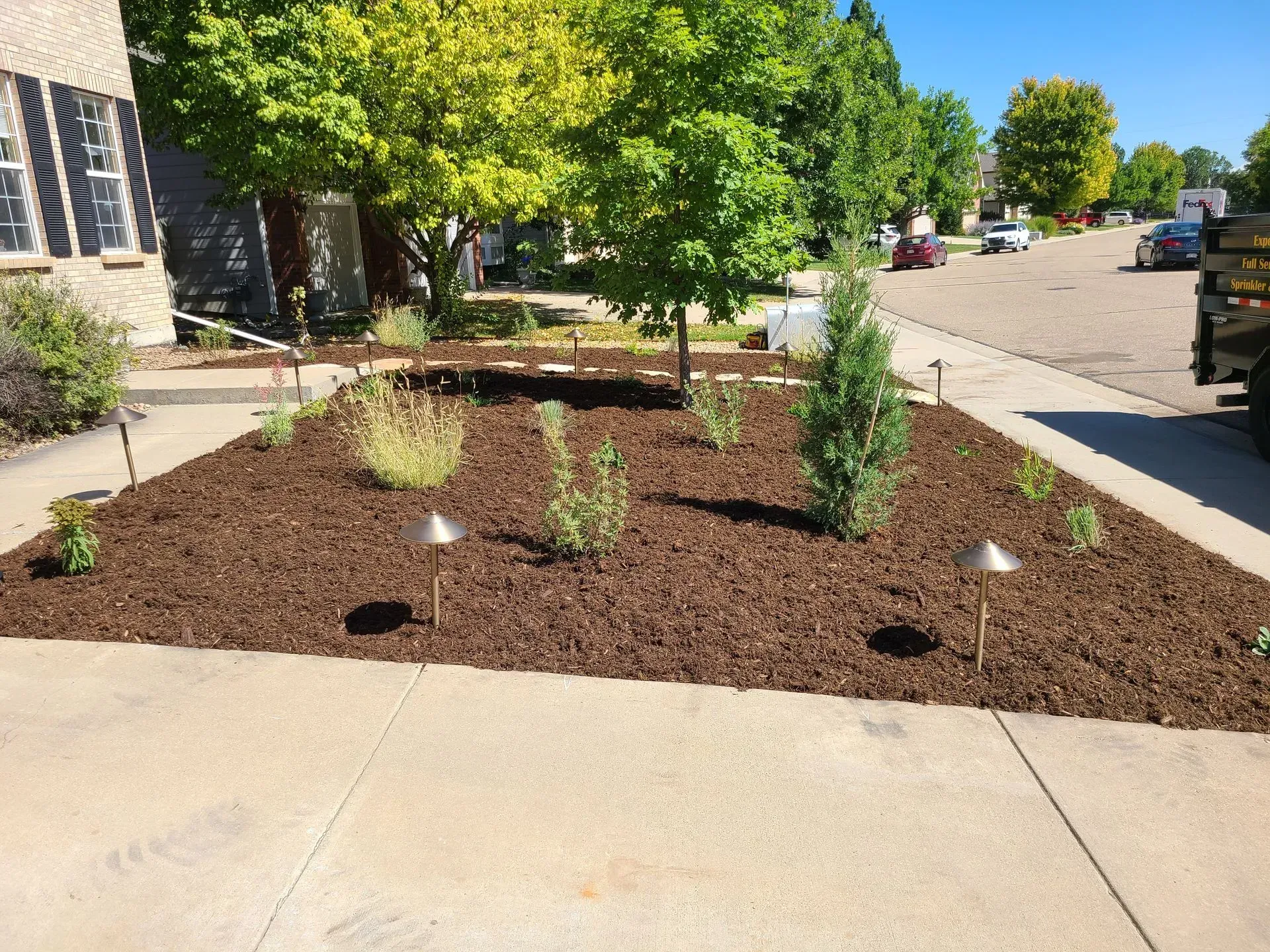 Garden bed with fresh mulch, newly planted greenery, and two path lights in front of a house on a sunny day.