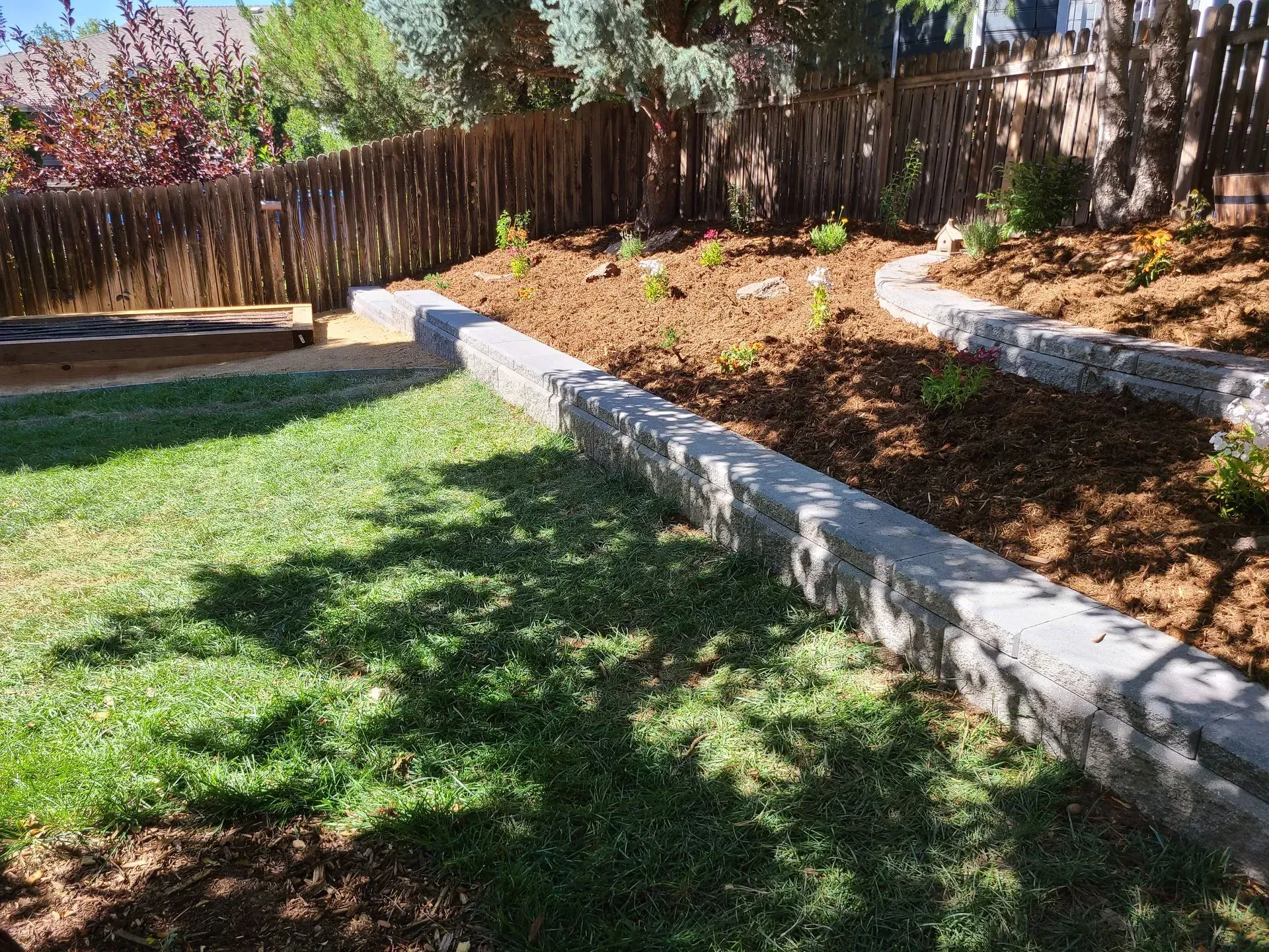 Retaining wall with tiered garden bed, filled with mulch and plants, grass lawn in foreground.