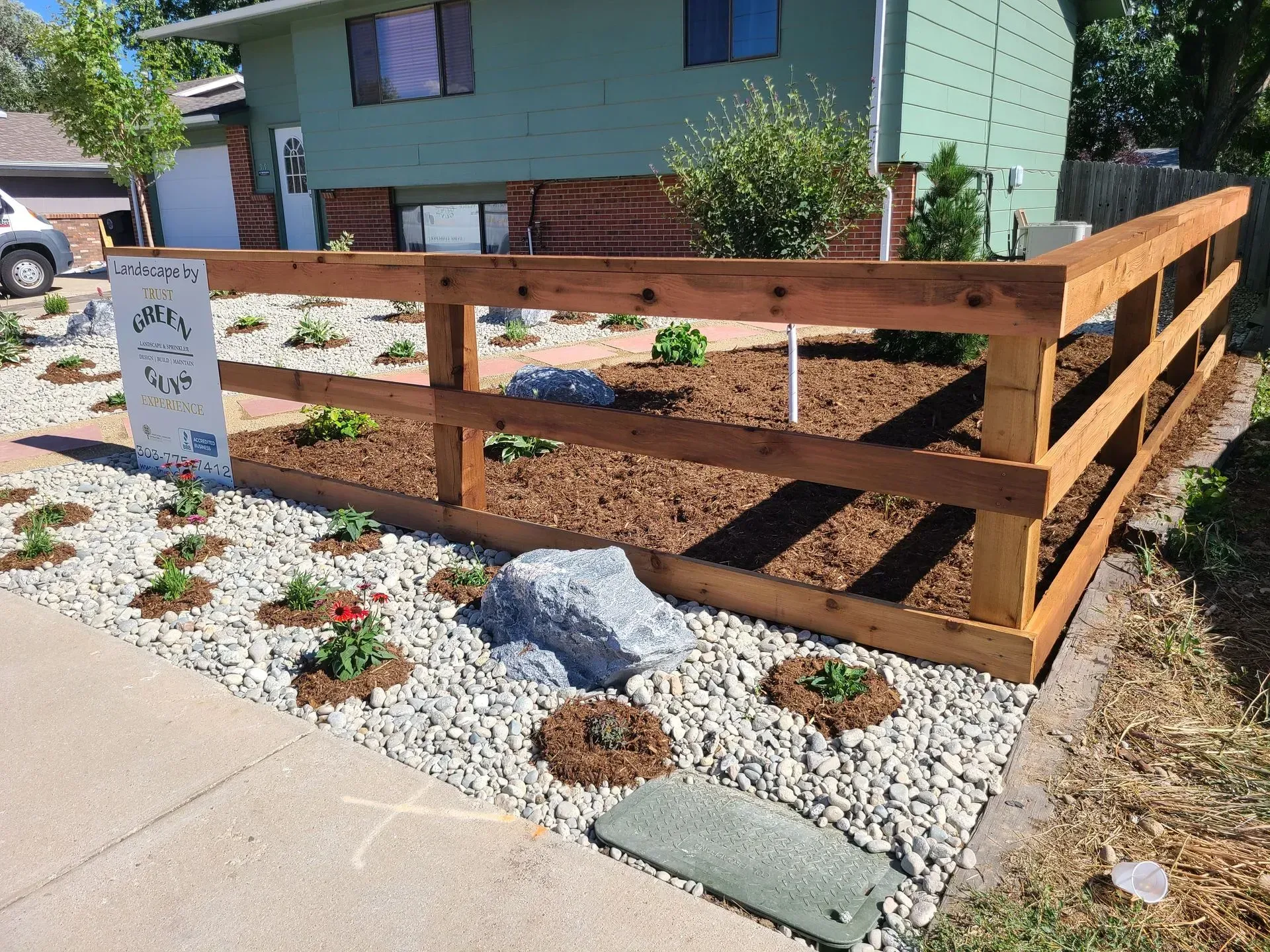 Wooden fence around a garden bed with plants, mulch, and rocks in front of a house.