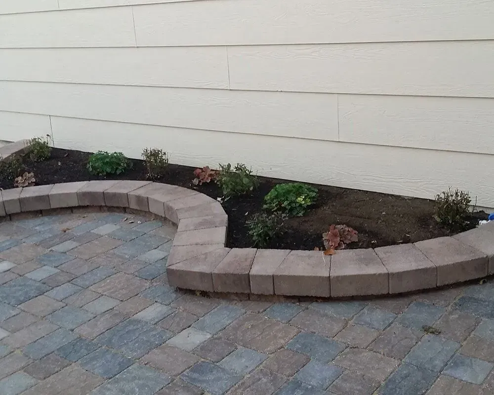 Raised garden bed with brick border, planted with small shrubs, against a beige house wall. Paved patio in front.