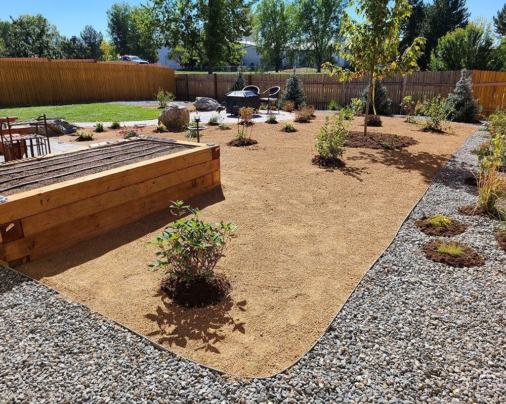 Backyard with gravel path, tan gravel bed, wood planter, plants, and fire pit near a wooden fence.