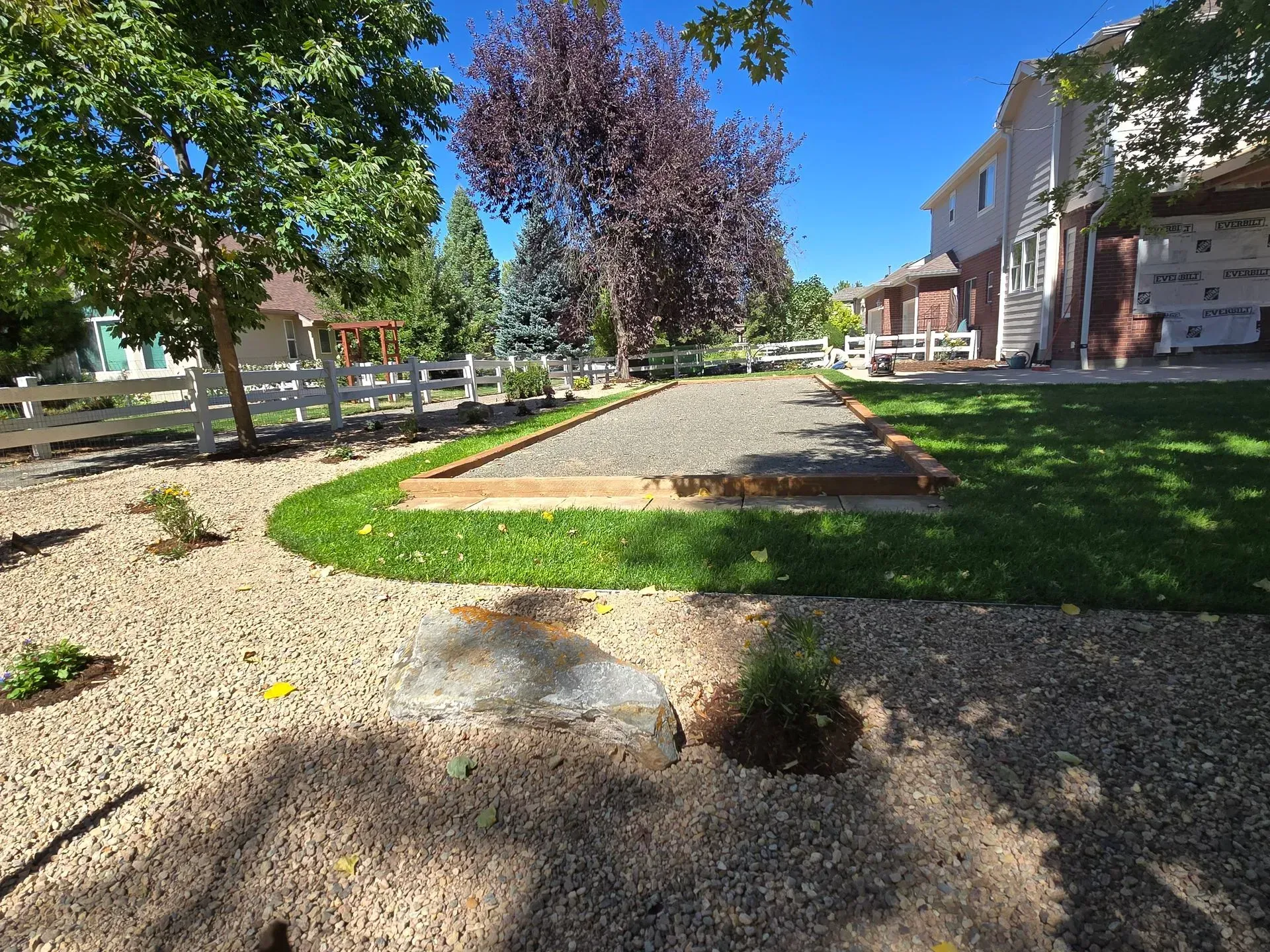 A gravel and grass backyard with a bocce ball court and white fence.