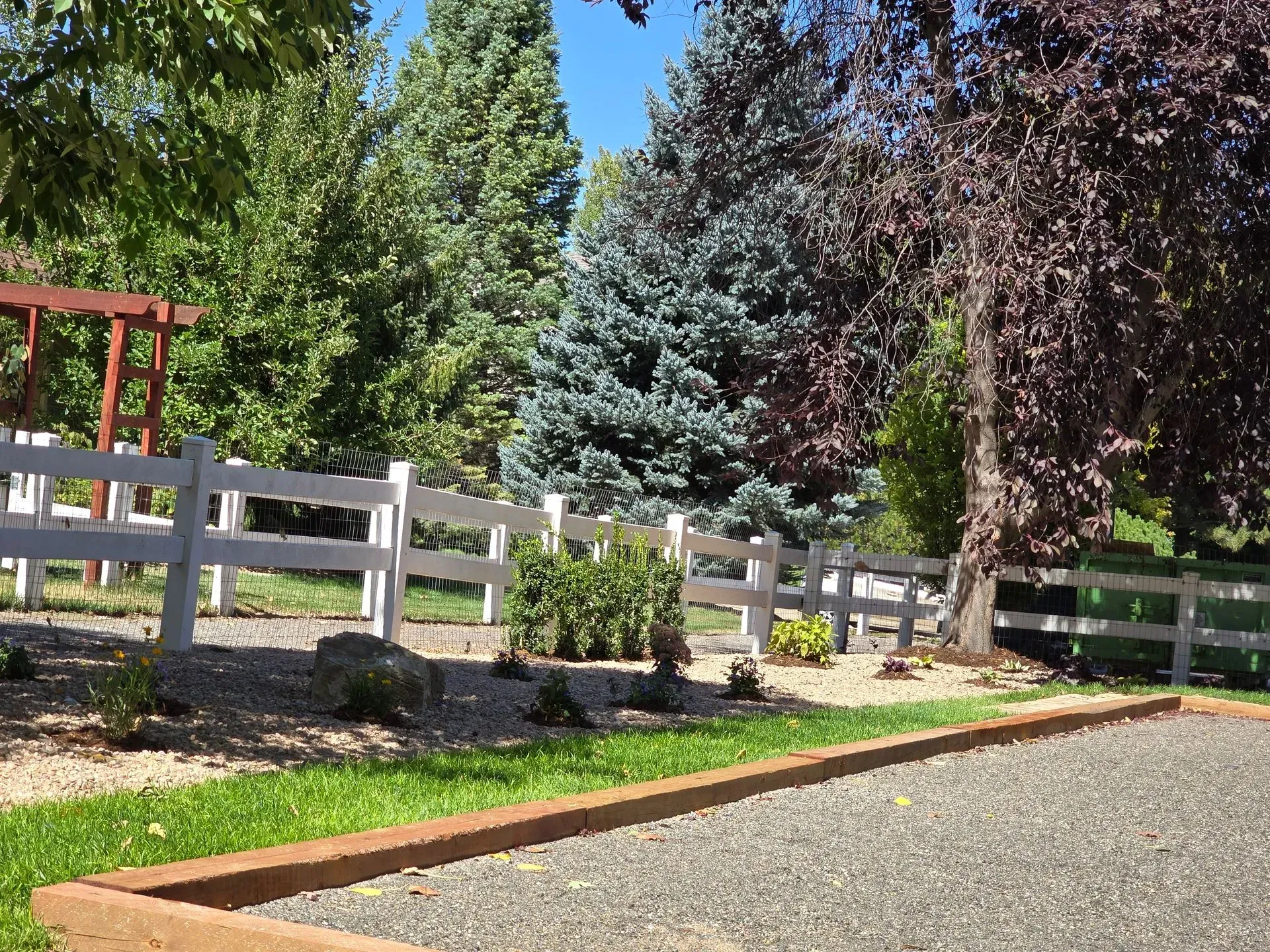 White fence in a yard with gravel and small plants, surrounded by trees.