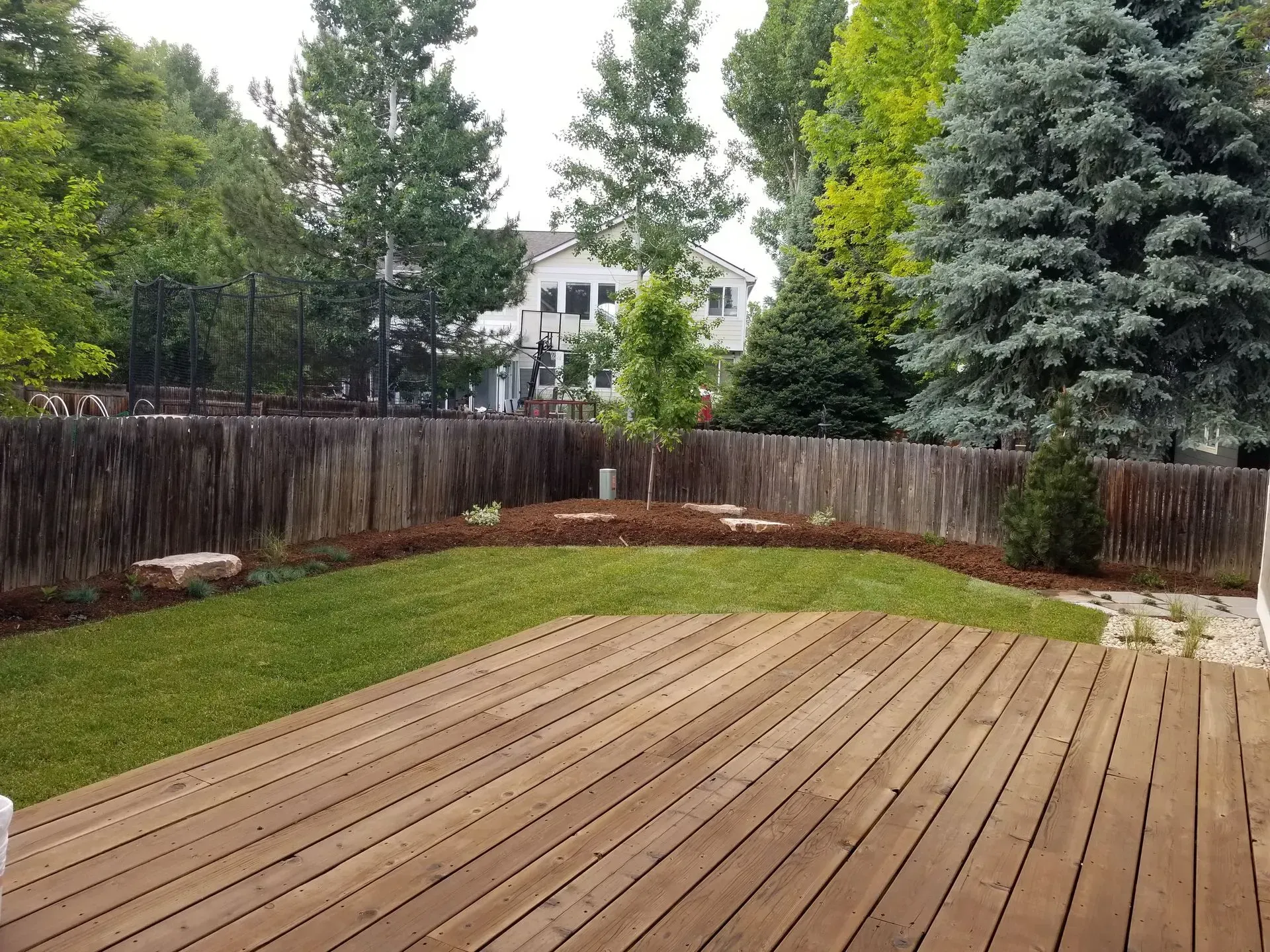 Wooden deck overlooks a backyard with grass, mulch, trees, and a wooden fence. A house is visible in the distance.