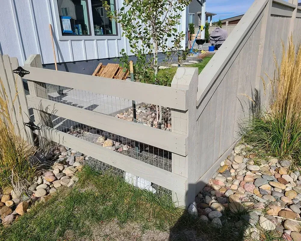 Beige wooden fence surrounding a recessed foundation area filled with rocks and grass.