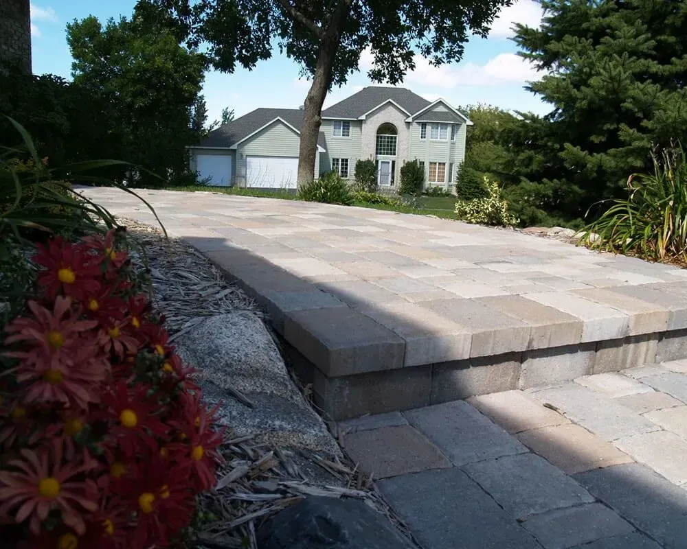 Stone steps lead to a paved driveway and large house with garage, surrounded by trees and flowers.