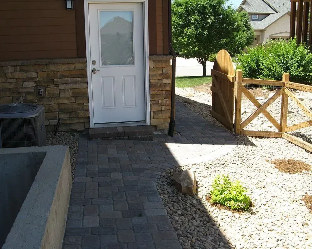 Paver walkway leading to a white door, with a stone wall and a wooden gate.