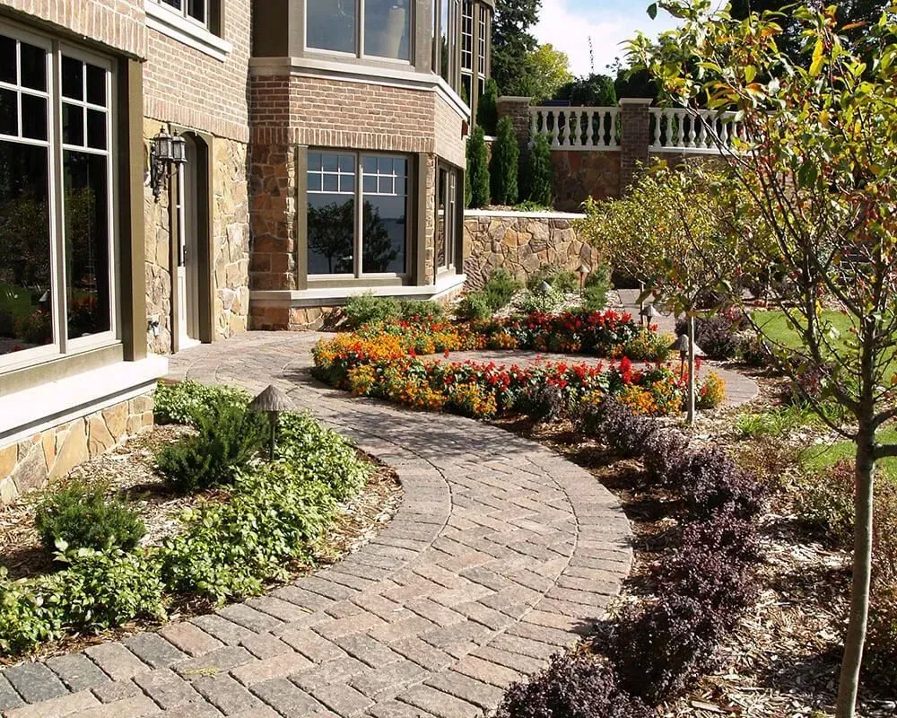 Brick pathway winds through landscaped garden near a brick and stone building.