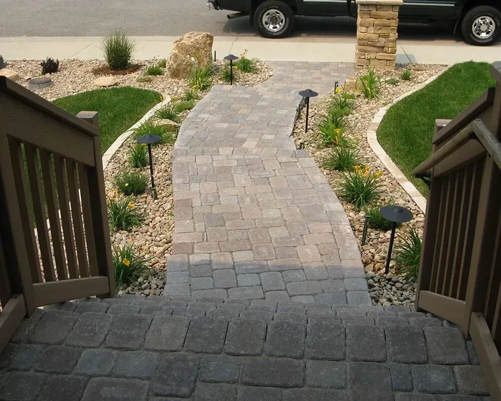 Stone pathway leading up to a porch with landscaping and lights, brown railings, and a vehicle in the background.
