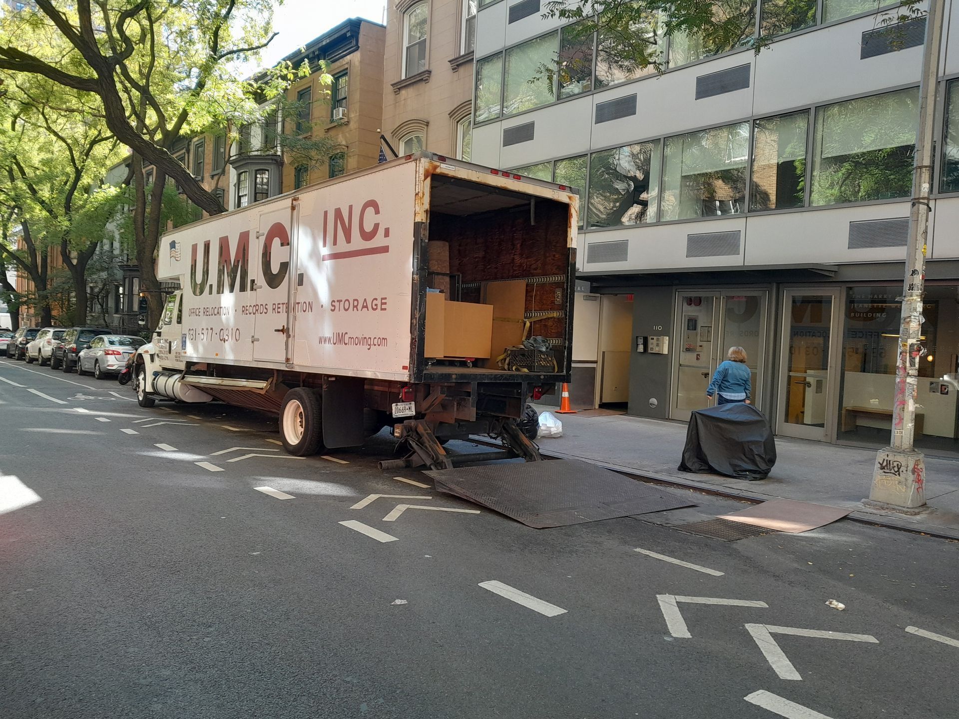Moving truck parked on a city street, loading boxes. U.M.C. INC. on the truck. Buildings and trees in the background.