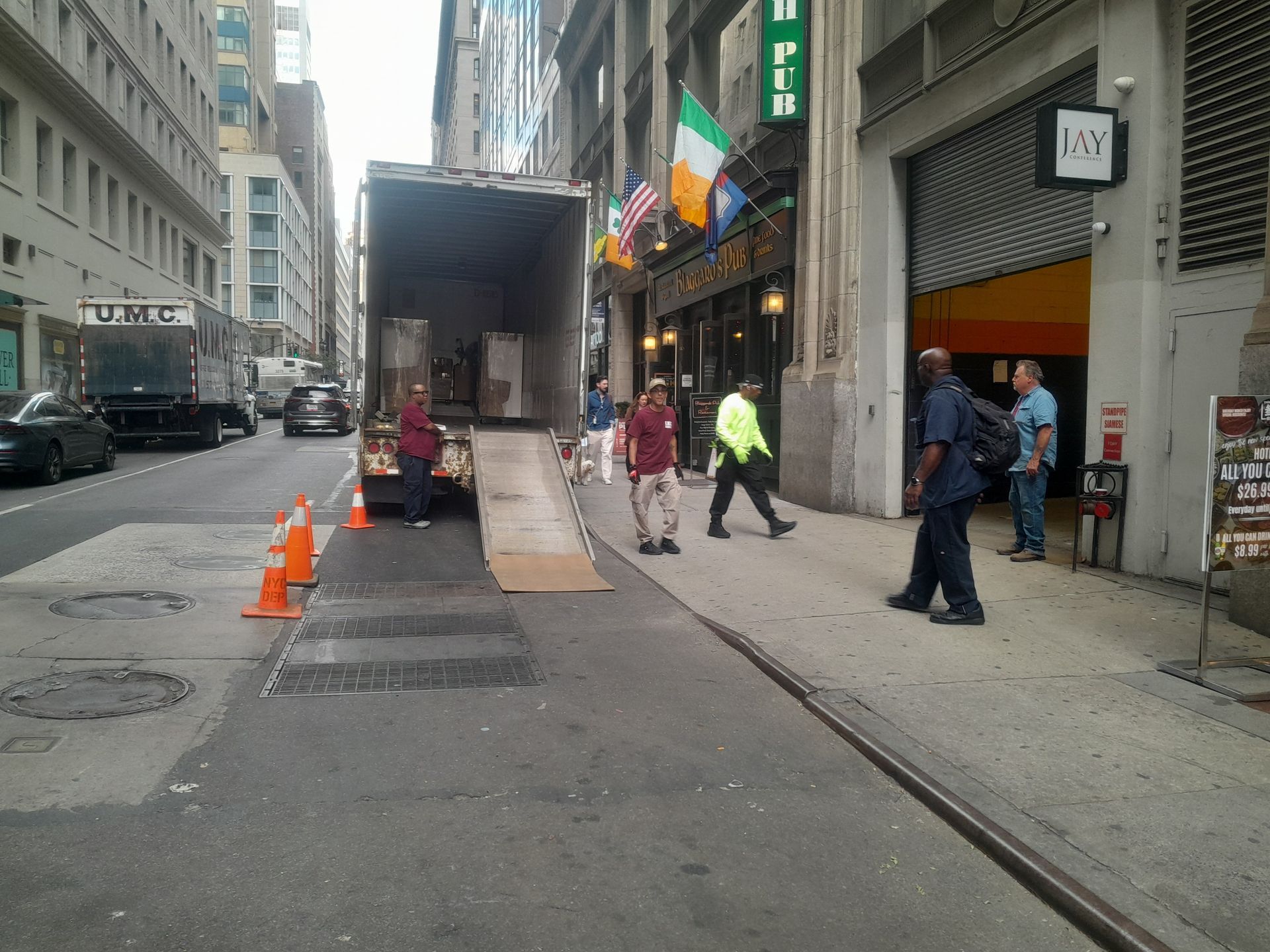 Moving truck on a city street with people loading items. Orange cones, a pub with flags visible.