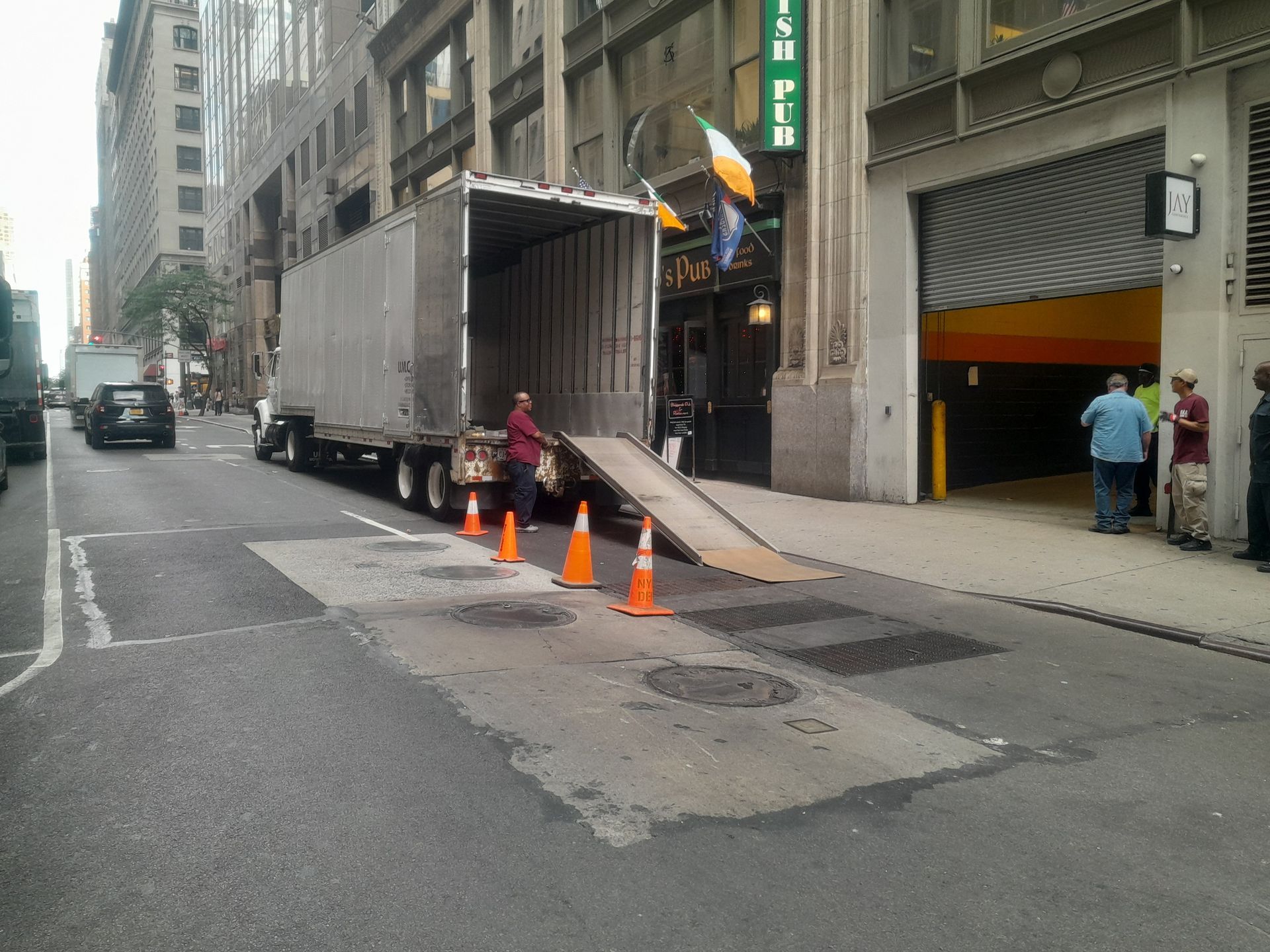 Truck unloading goods on city street in front of an Irish pub and parking garage. Cones and people present.