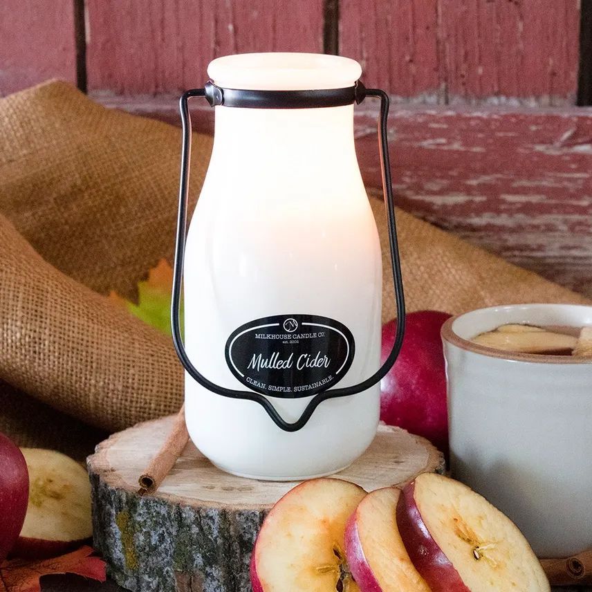 A glowing candle in a white milk jug with a black handle, surrounded by apples and cider.