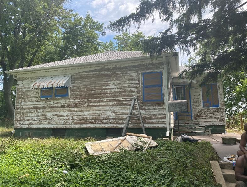 Old house with peeling paint, blue-taped windows, ladder, and overgrown yard.