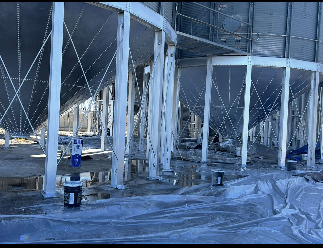 Grain storage silos with white support beams, under construction; blue paint buckets present.
