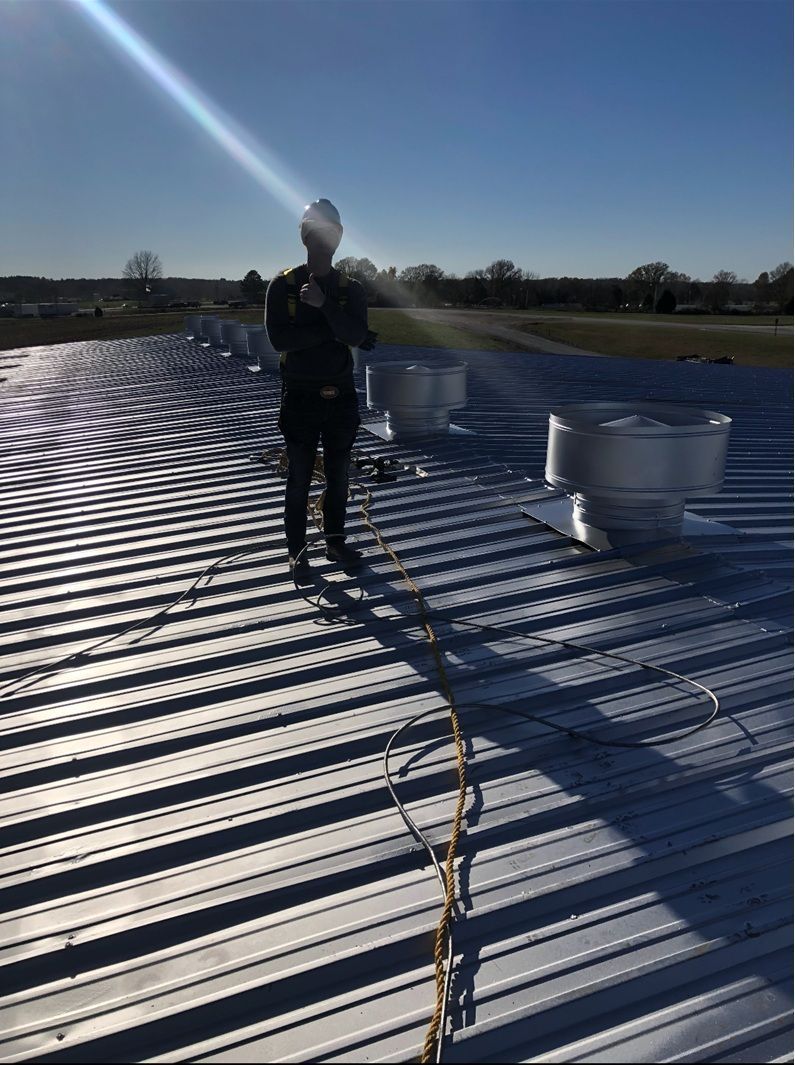 Person on a corrugated metal roof wearing a harness, working near vents on a sunny day.