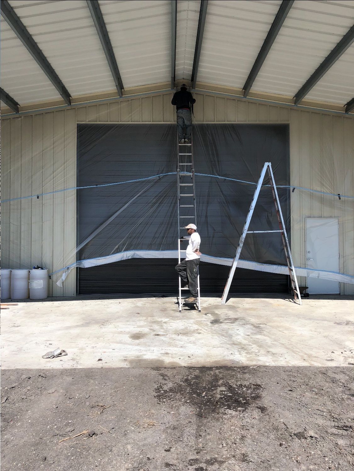 Two people on ladders working on a building's overhead door. One person is on the roof. Gray concrete.
