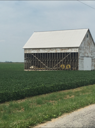White weathered barn with open side in a green field, under a cloudy sky.