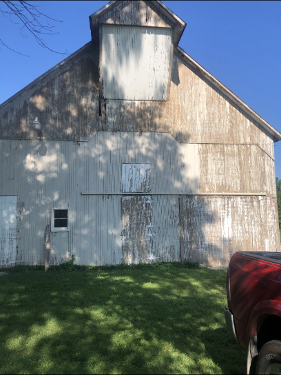 A weathered barn with a faded white exterior, a small window, and a red vehicle visible on the right.