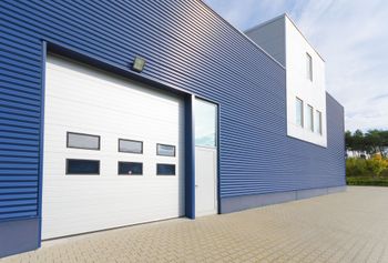 Blue corrugated building with a large garage door, a side door, and a white section with windows.