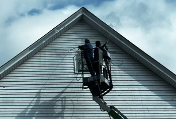 Two workers in a lift, working on the siding of a white house under a cloudy sky.
