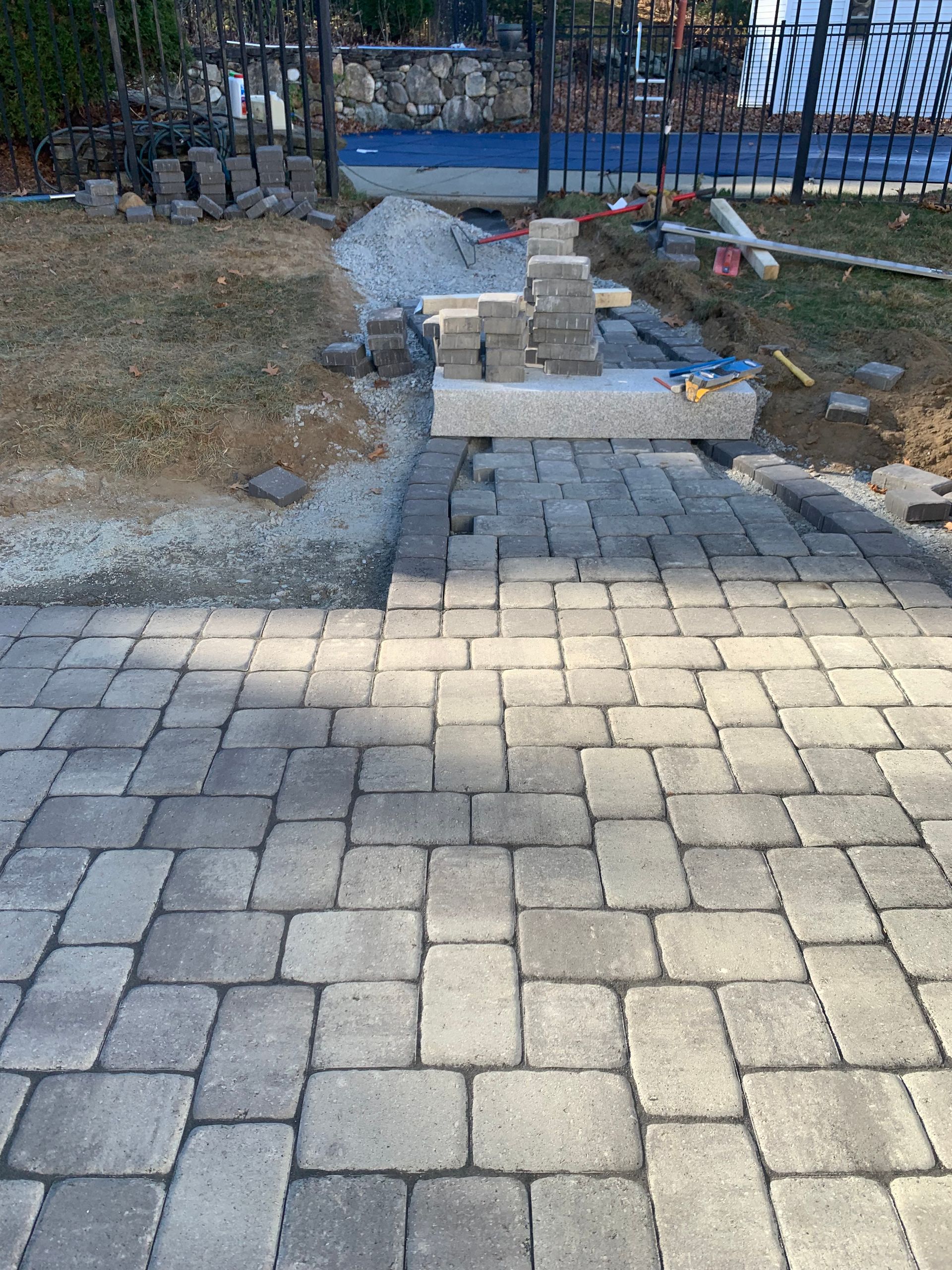 Pathway construction with gray pavers, gravel, and blocks near a fenced pool.