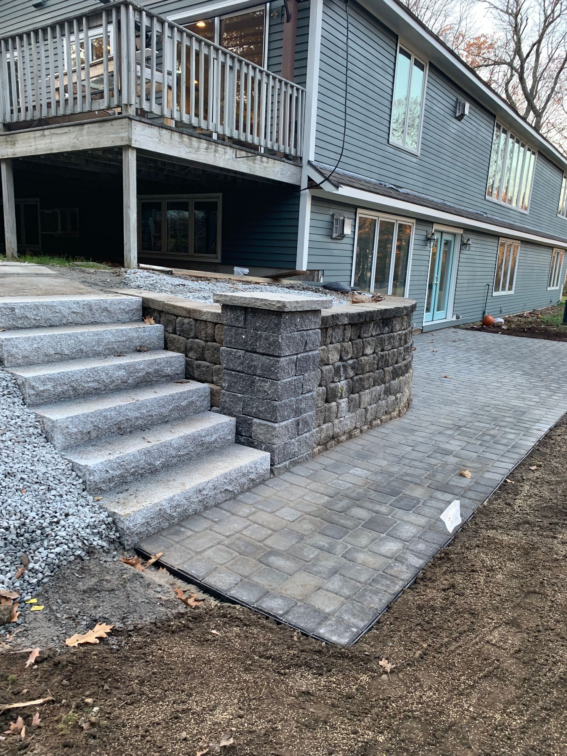 Stone steps lead to a patio with a retaining wall next to a two-story blue house.