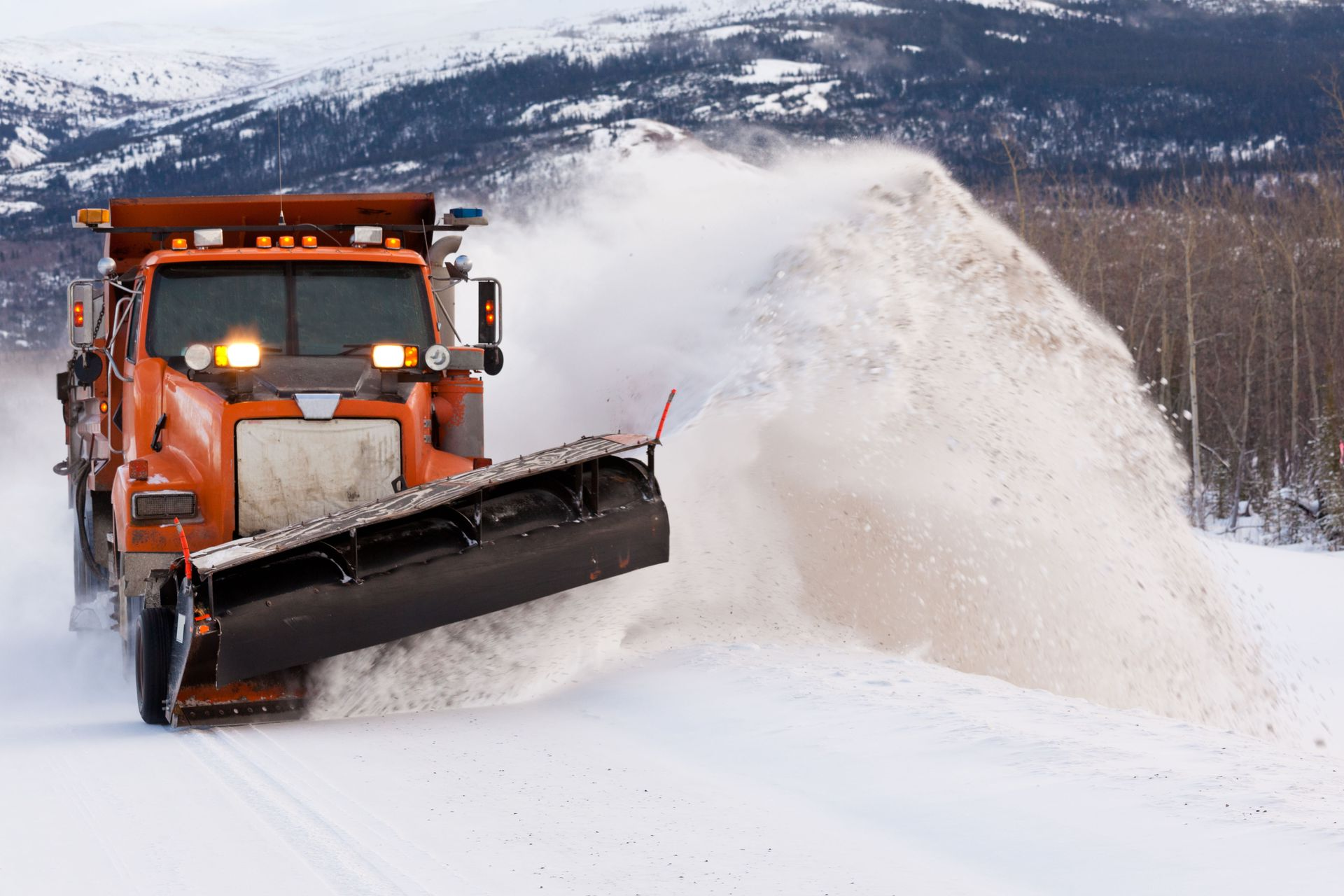 Orange snowplow clearing snow on a road with snowy mountains and trees in the background.