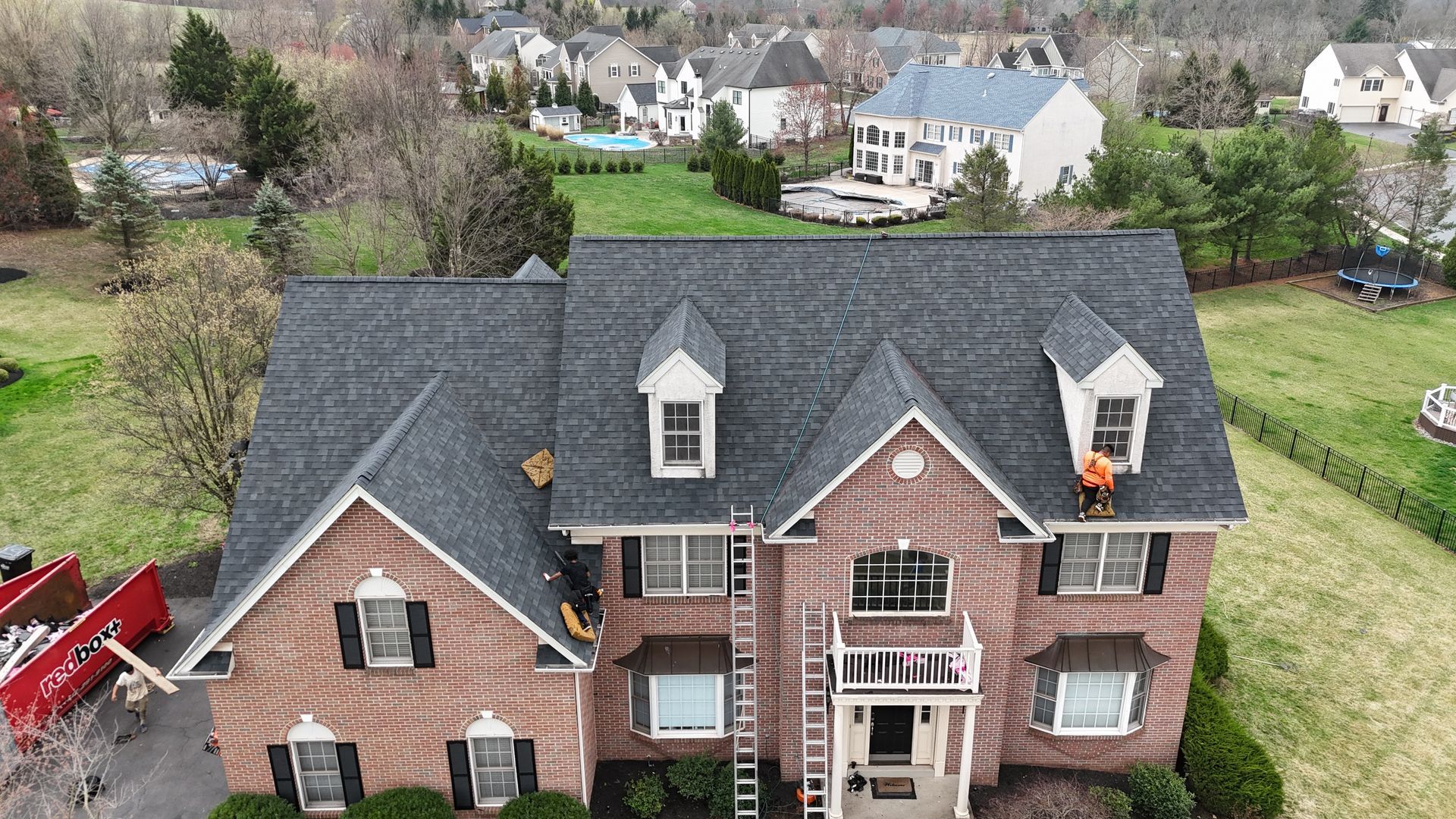 Brick house with new dark roof; workers on roof; houses in background.