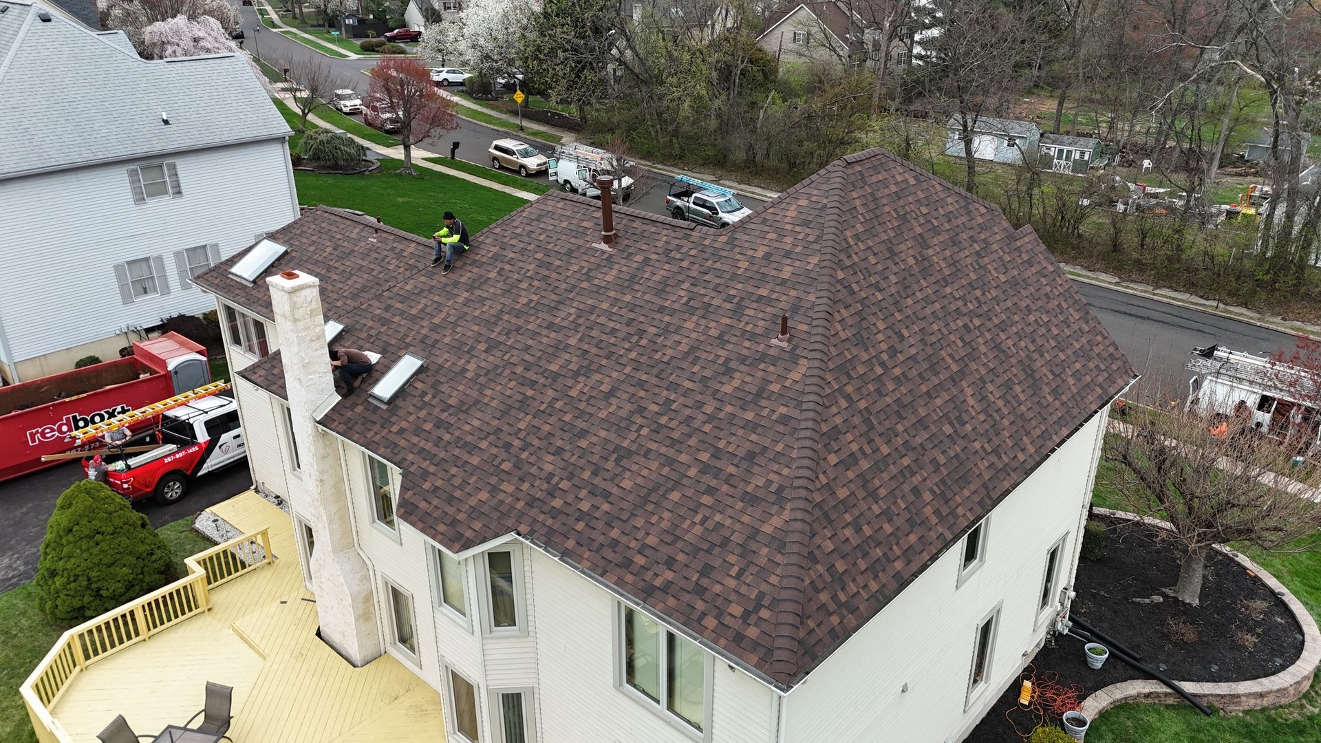 A roofer on a brown shingle roof. White house with wooden deck, red truck parked in front.