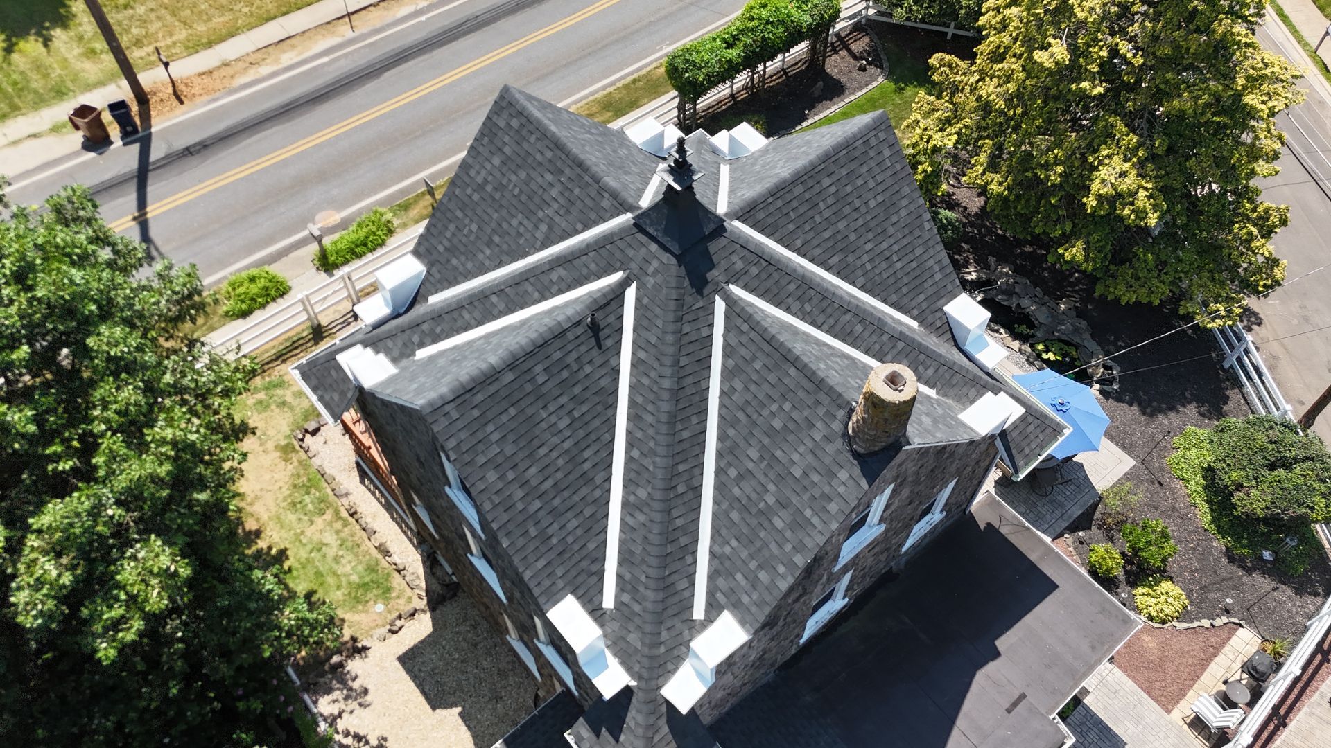 Overhead view of house with dark roof and white accents. Street and trees surround the house.