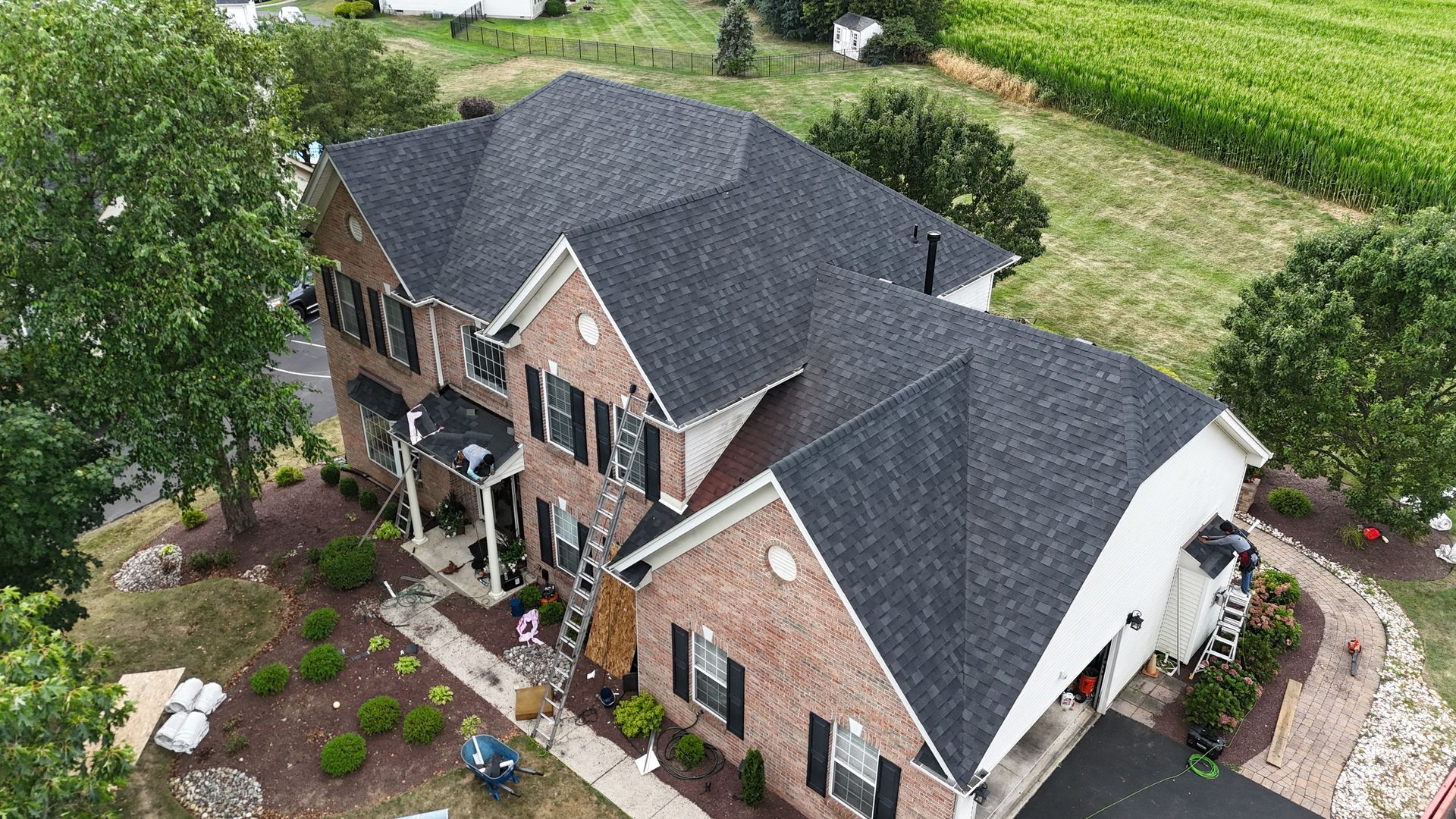 Two-story brick house with a dark roof, black shutters, and a paved driveway surrounded by green grass.