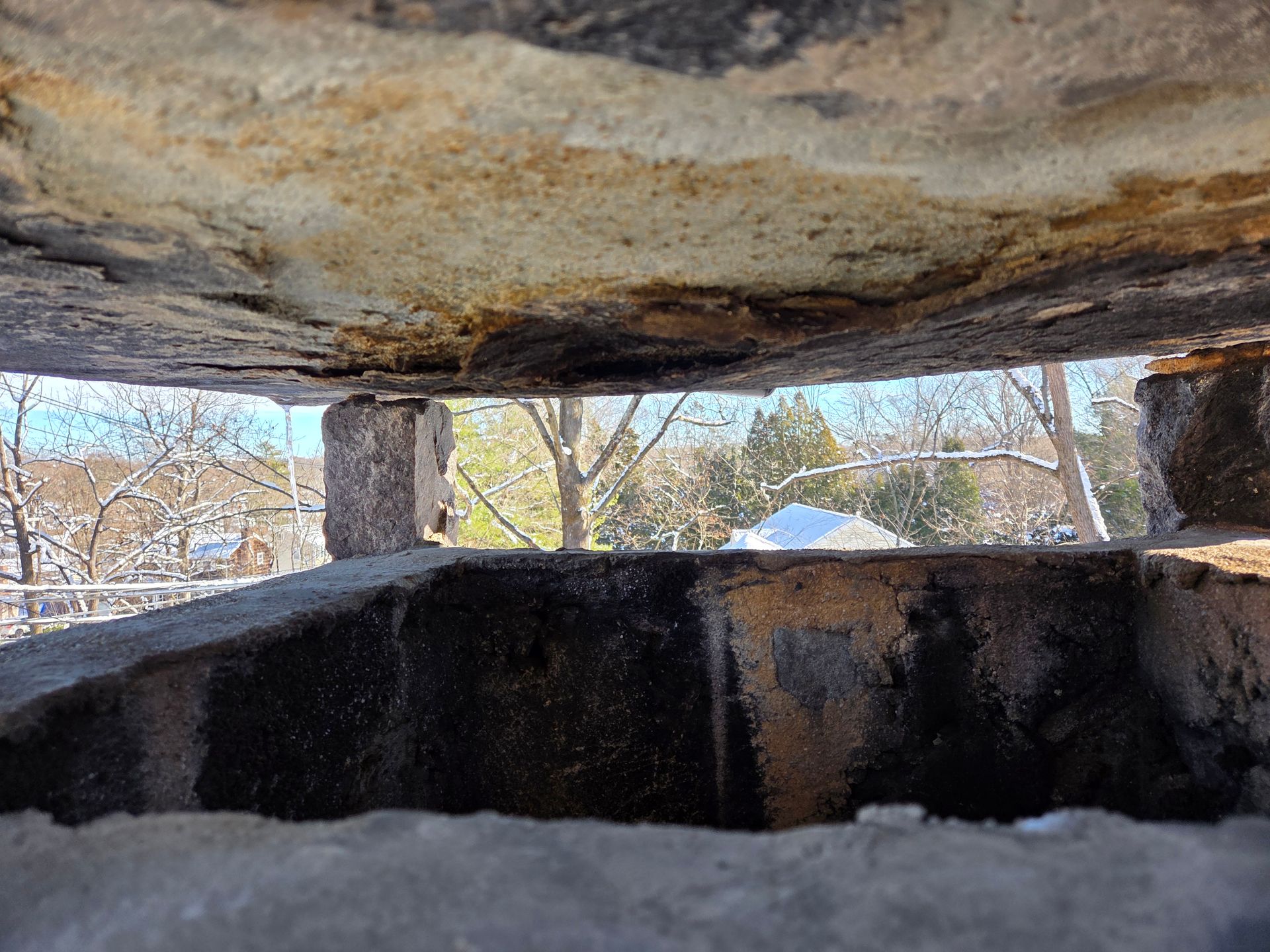 Looking out from a stone structure's opening, trees and a building are visible against a bright sky.