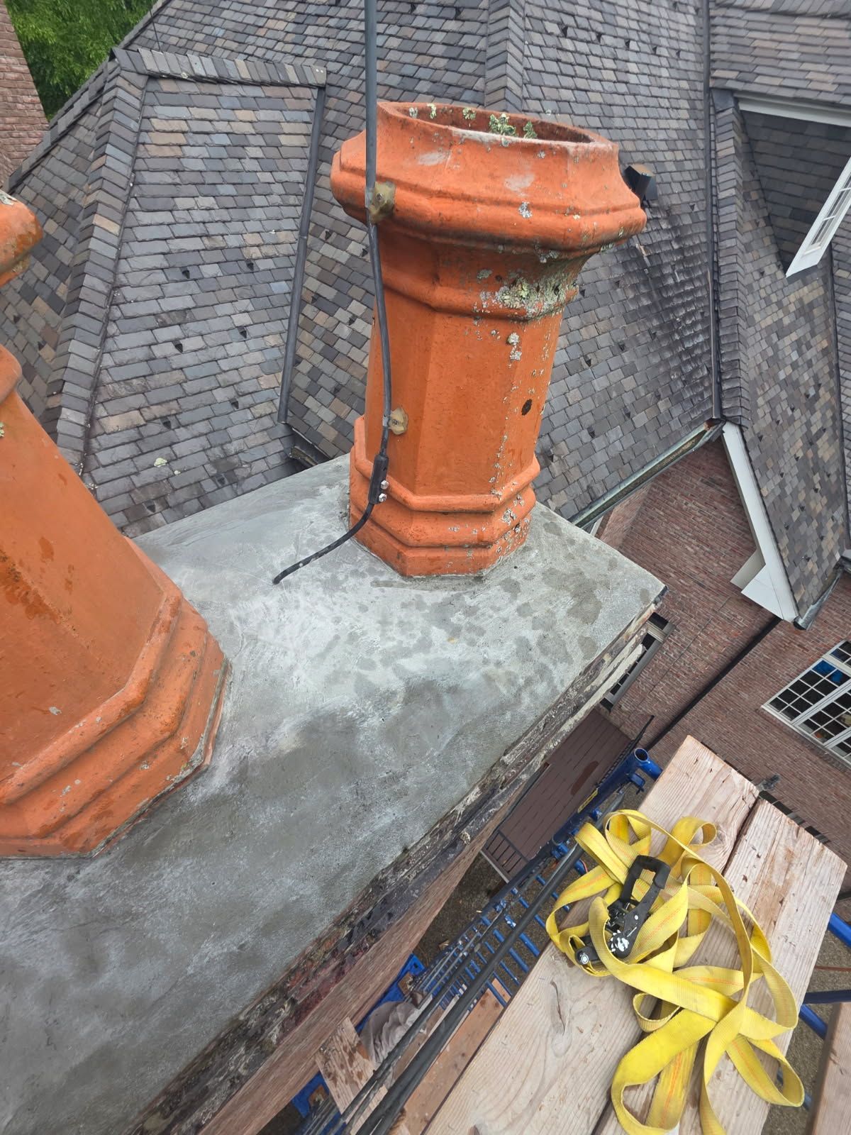 Damaged brick chimney corner with missing bricks, near gray flashing, scaffolding, and green grass.