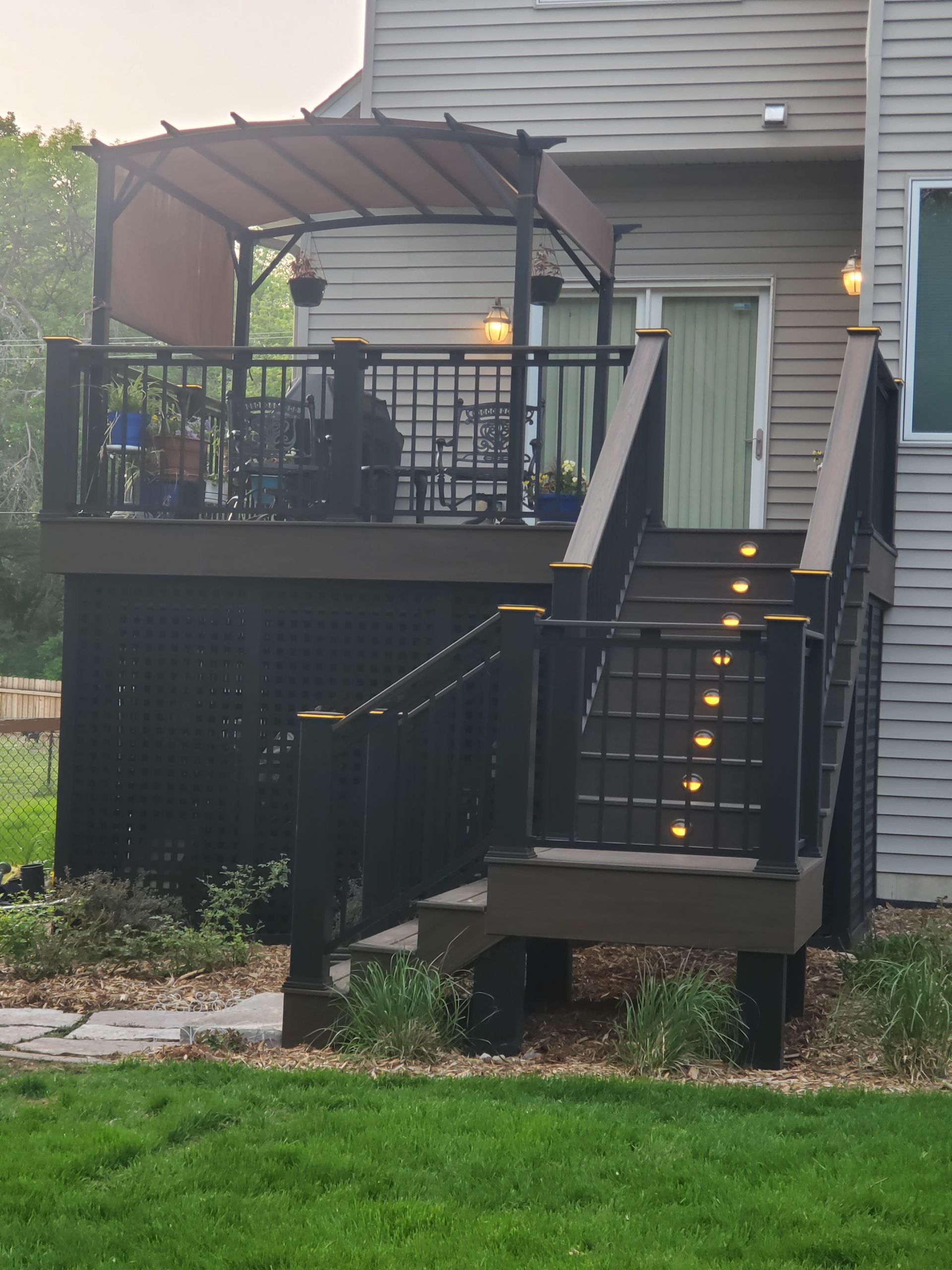 An elevated deck with a brown shade canopy and a staircase with illuminated risers, attached to the back of a house.