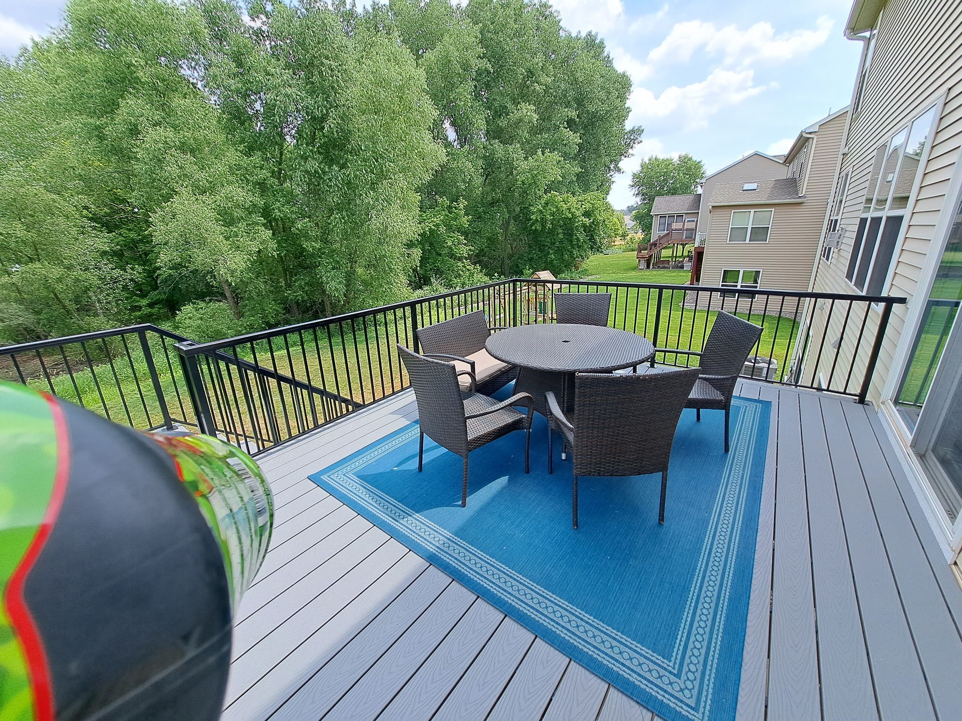 A gray deck with a blue patterned rug, a round table, and four wicker chairs, overlooking a lush green yard.