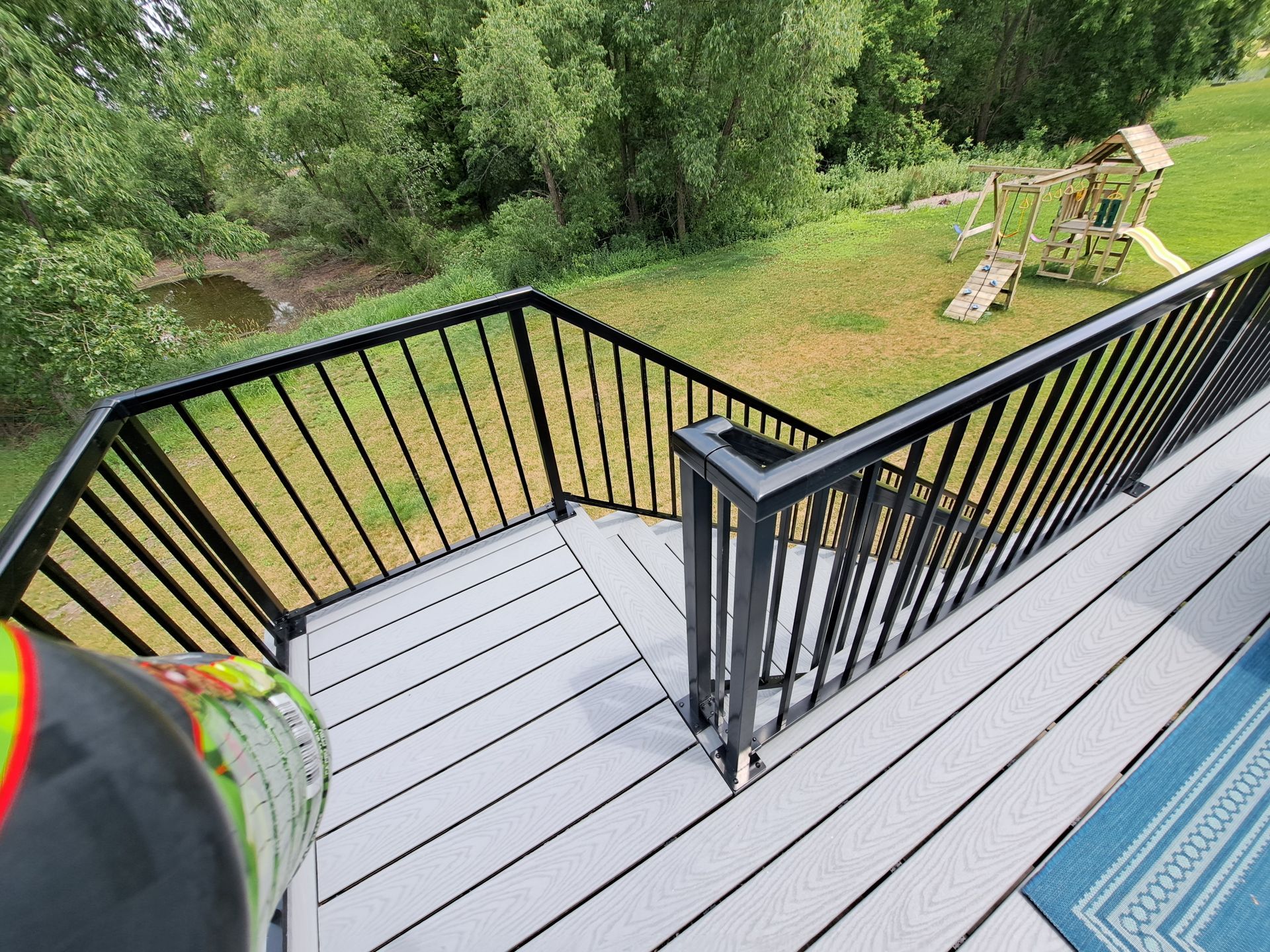 A view from a grey composite deck, overlooking a grassy backyard with a wooden playset, framed by black metal railings.