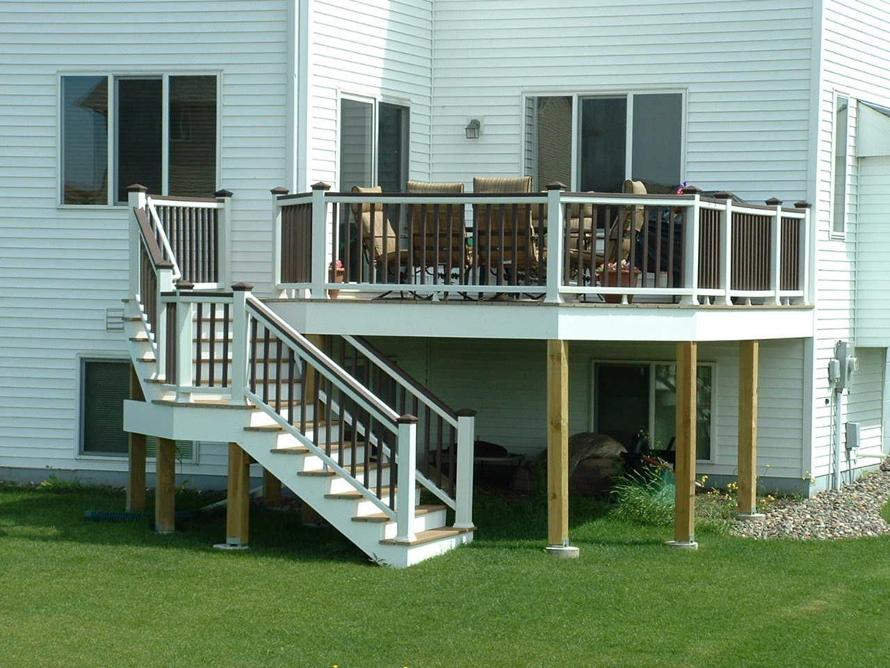 A white house with a raised wooden deck featuring dark brown railings, steps leading down, and outdoor patio furniture.