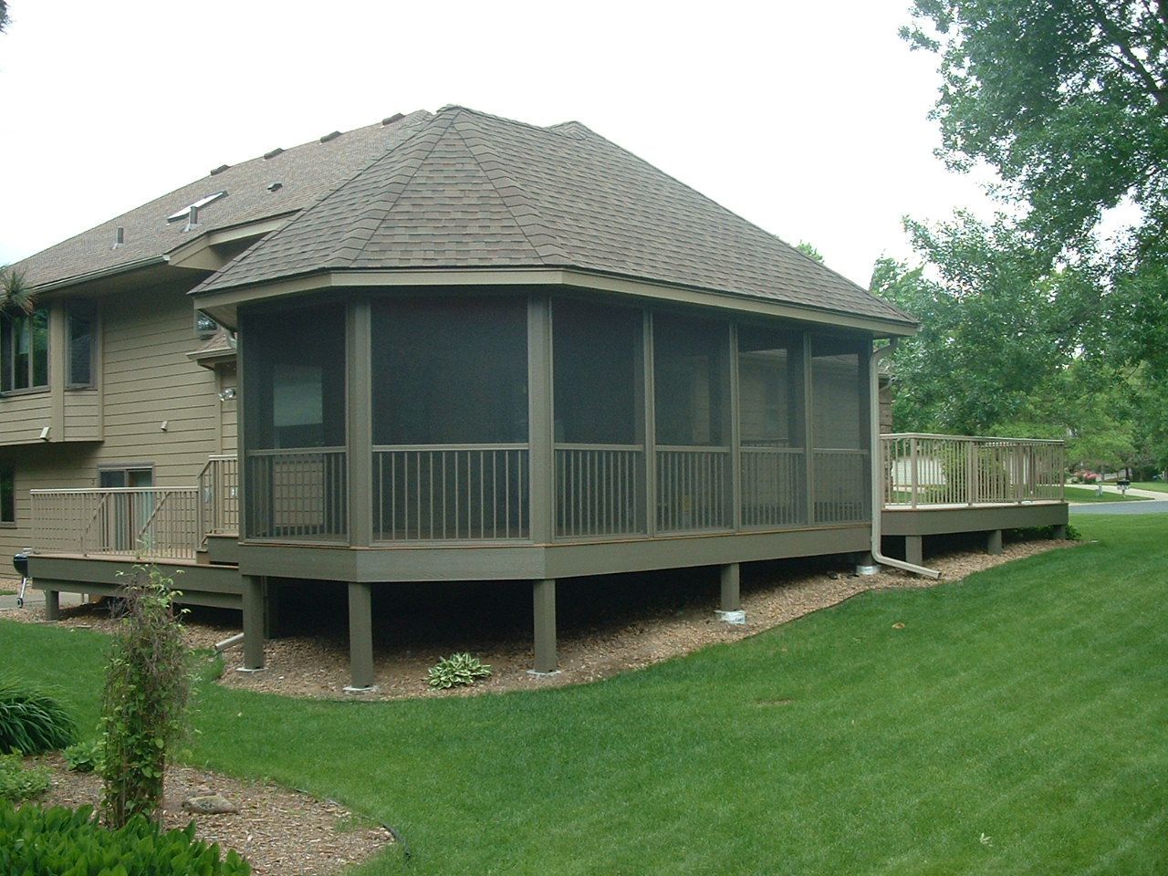 A tan house featuring a screened-in porch with a gray roof and a wooden deck extending into a green grassy yard.